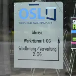 Hinweisschild zu Mensa und Schulleitung hinter einer Glastür, daneben ein Plakat für das Schulfest
