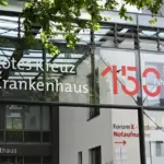 Gläserner Haupteingang des Rotes Kreuz Krankenhauses mit Hinweis zur Notaufnahme