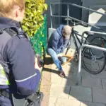 Ein Polizist von hinten vor mehreren an einem Metallständer angebundenen Fahrrädern, im Hintergrund ein modernes Bürogebäude.