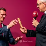 Zwei Männer in Anzügen überreichen sich gegenseitig einen ornaten Stab vor Bundesrat-Branding.